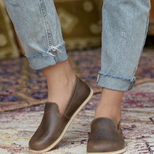 May include: A pair of brown leather slip-on shoes with a cream-colored sole. The shoes are worn by a person wearing blue jeans.