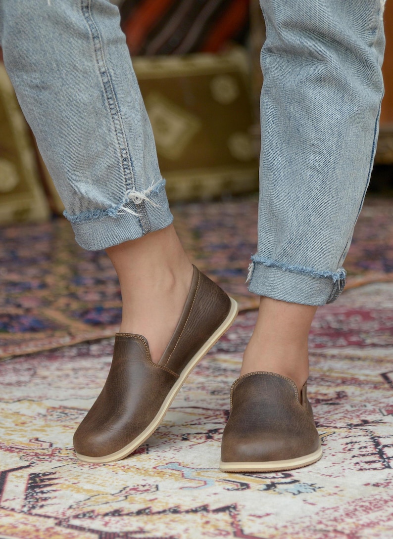 May include: A pair of brown leather slip-on shoes with a cream-colored sole. The shoes are worn by a person wearing blue jeans.
