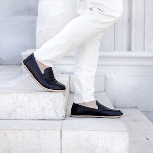 Op de afbeelding: Een paar zwarte leren loafers met een cr&egrave;mekleurige zool. De loafers worden gedragen door iemand die een witte broek draagt en op een trap staat.