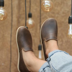 May include: A pair of brown leather slip-on shoes with white soles. The shoes are worn by a person sitting with their legs crossed, wearing blue jeans.