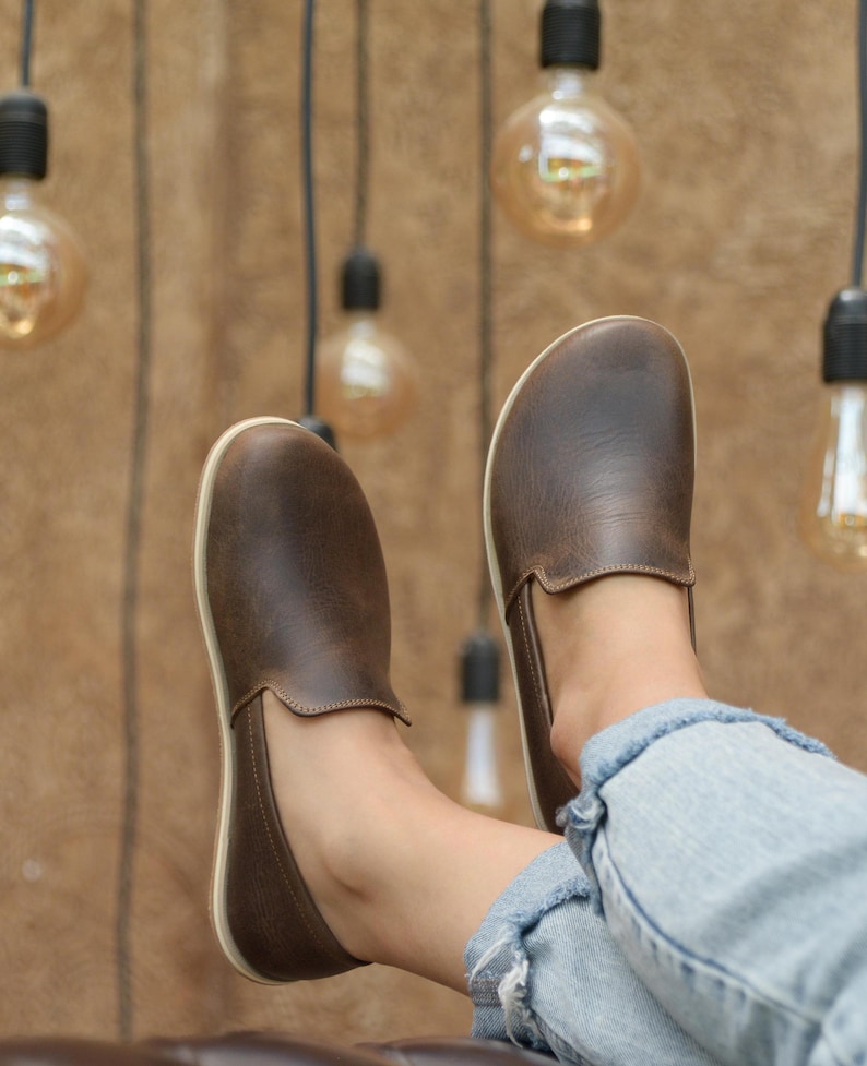 May include: A pair of brown leather slip-on shoes with white soles. The shoes are worn by a person sitting with their legs crossed, wearing blue jeans.