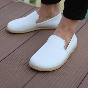 May include: A pair of white leather slip-on shoes with a light brown rubber sole. The shoes are worn by a person standing on a wooden surface.
