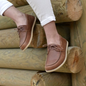 May include: A pair of brown leather boat shoes with laces. The shoes are worn by a person sitting on a wooden log.