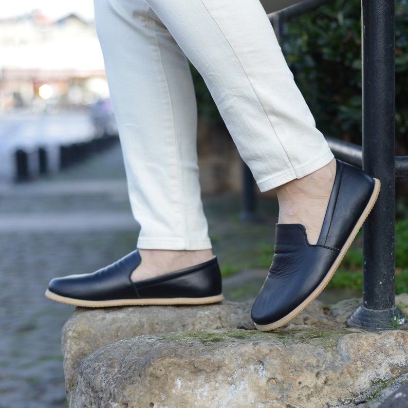 May include: Black leather slip-on shoes with a tan sole. The shoes are worn with cream-colored pants. The shoes have a minimalist design and are suitable for casual wear. The shoes are shown outdoors.