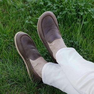 May include: A pair of brown leather loafers with a cream colored sole. The loafers are worn by a person wearing white pants.