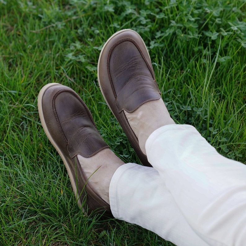 May include: A pair of brown leather loafers with a cream colored sole. The loafers are worn by a person wearing white pants.