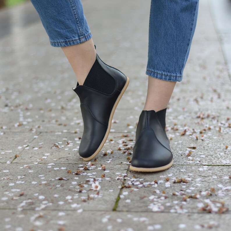 May include: Black ankle boots with a minimalist design. The boots have a smooth leather upper, elastic side panels, and a light-colored sole. The boots are worn with blue jeans. The boots are approximately 6 inches in height.