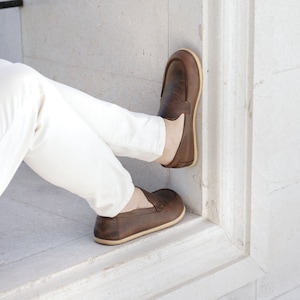 May include: A pair of brown leather slip-on shoes with a light brown sole. The shoes are worn by a person sitting on a white stone surface.