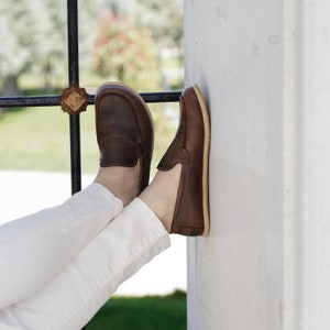 May include: A pair of brown leather loafers with white stitching. The loafers are worn by a person wearing white pants.