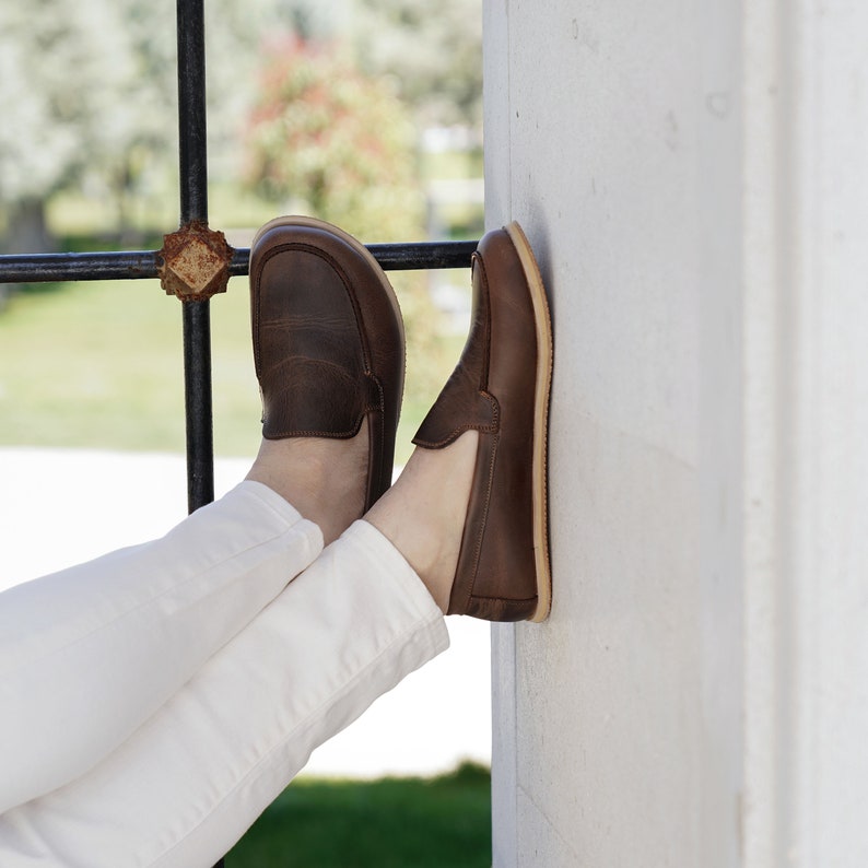 May include: A pair of brown leather loafers with white stitching. The loafers are worn by a person wearing white pants.