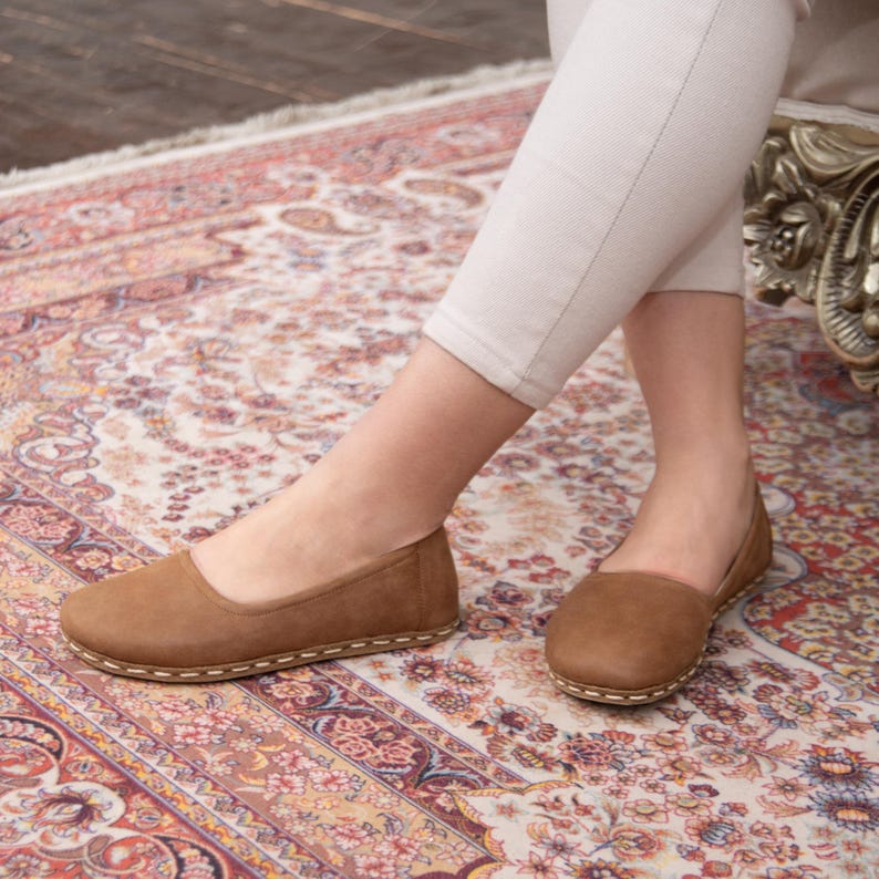 May include: A pair of brown leather ballet flats with a stitched edge. The flats are on a patterned rug.
