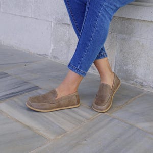May include: A pair of brown suede loafers with a rounded toe and a light-colored sole. The shoes are worn with blue jeans. The loafers have a classic slip-on design and are made of a soft, textured material.