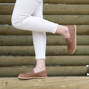 May include: A pair of brown leather boat shoes with laces. The shoes are worn by a person standing on a wooden surface.