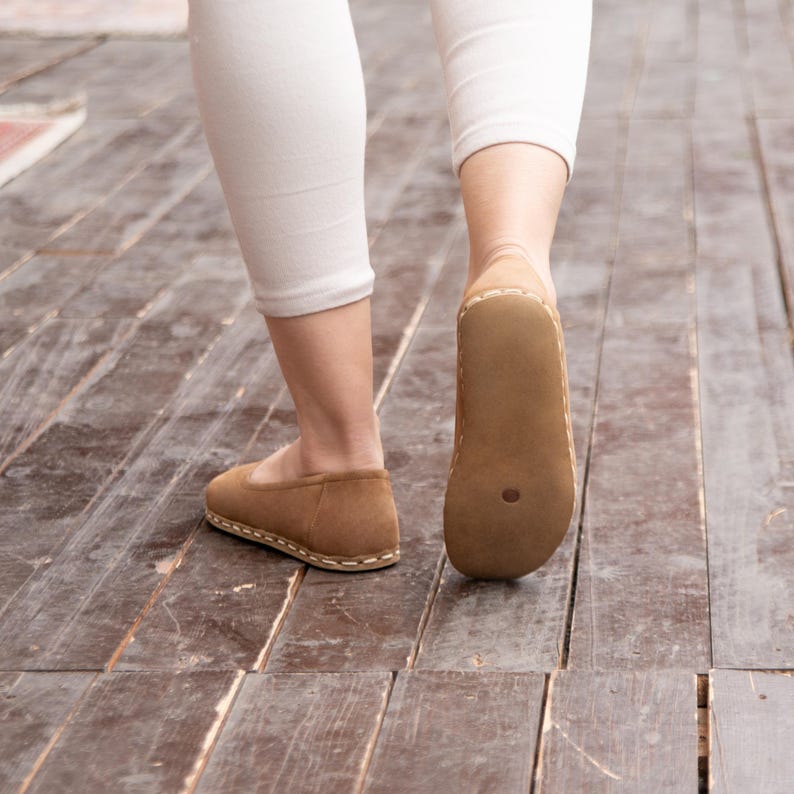 May include: A pair of brown leather slip-on shoes with a stitched seam around the edge. The shoes are worn by a person walking on a wooden floor.