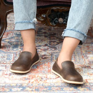 May include: A pair of brown leather slip-on shoes with a white sole. The shoes are worn by a person sitting on a chair with their legs crossed.