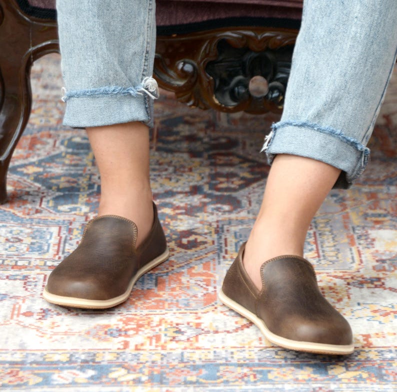 May include: A pair of brown leather slip-on shoes with a white sole. The shoes are worn by a person sitting on a chair with their legs crossed.
