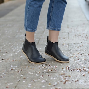 Puede incluir: Botas Chelsea de cuero negro con paneles laterales elásticos y suela de color claro. Las botas se llevan con vaqueros azules. La imagen está tomada al aire libre en una acera de piedra.