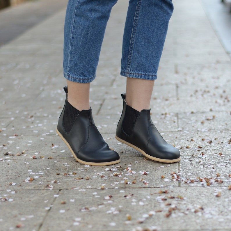 May include: Black leather Chelsea boots with elastic side panels and a light-colored sole. The boots are worn with blue jeans. The image is taken outdoors on a stone walkway.