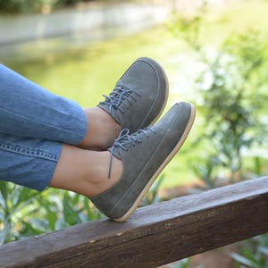 Può includere: Un paio di scarpe in pelle scamosciata grigie con lacci e suola marrone chiaro. Le scarpe sono indossate da una persona seduta su un corrimano in legno.
