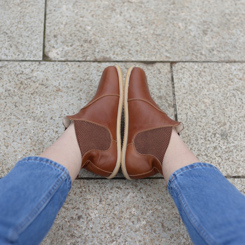 May include: A pair of brown leather ankle boots with a mesh panel on the side. The boots are worn by a person wearing blue jeans.