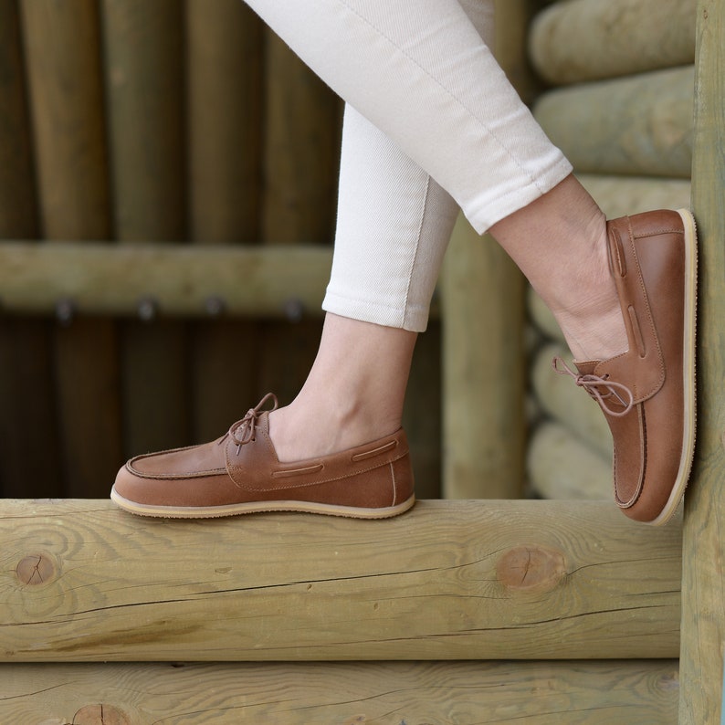 May include: A pair of brown leather boat shoes with laces. The shoes are worn by a person wearing white pants and are resting on a wooden surface.