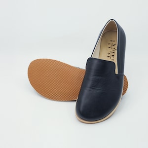 May include: A pair of black leather slip-on shoes with a rounded toe and a light brown sole. The sole of one shoe is visible, showing a textured pattern. The interior of the shoe is a light color, and the brand name is printed on the side.