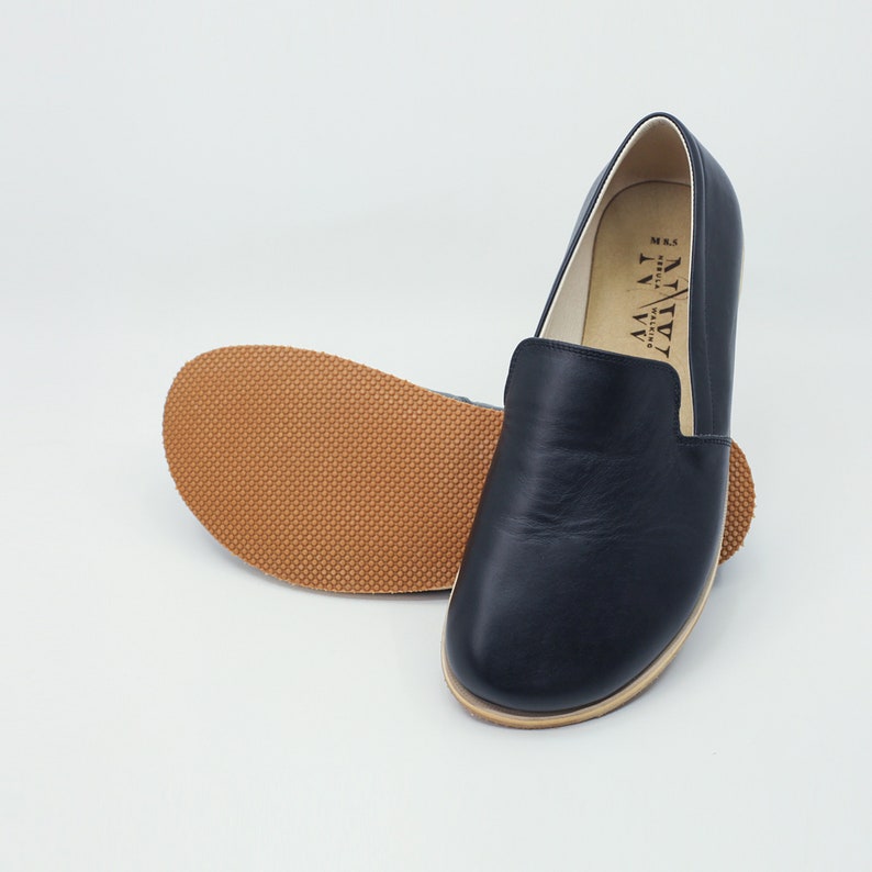 May include: A pair of black leather slip-on shoes with a rounded toe and a light brown sole. The sole of one shoe is visible, showing a textured pattern. The interior of the shoe is a light color, and the brand name is printed on the side.