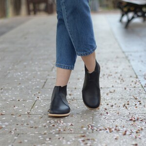 May include: Black ankle boots with a low heel and a rounded toe. The boots have a smooth leather-like appearance and a light-colored sole. The person is wearing blue jeans. The boots are suitable for everyday wear.