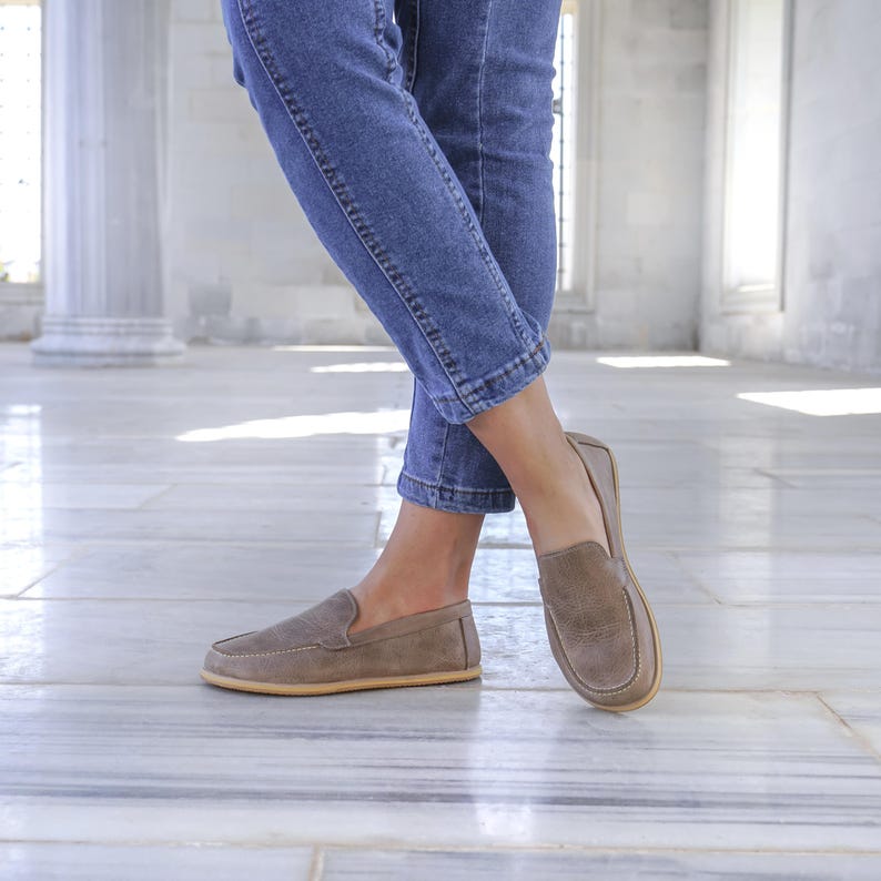 May include: A pair of taupe suede slip-on loafers with a light brown sole. The shoes are worn with blue denim jeans. The image is taken indoors with natural light.