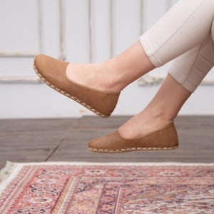 May include: A pair of brown leather ballet flats with a stitched edge. The flats are worn by a person sitting on a rug.