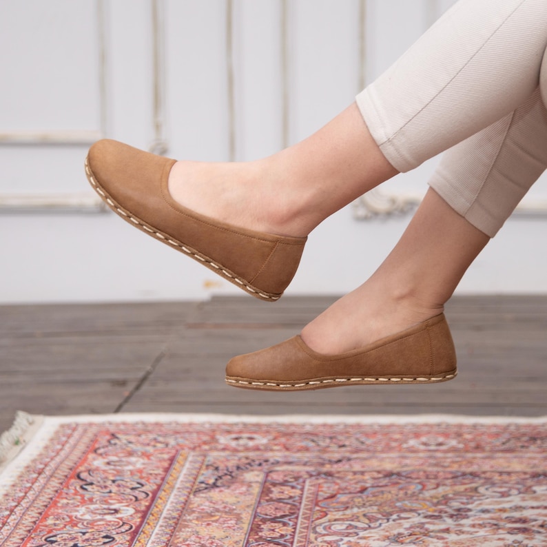 May include: A pair of brown leather ballet flats with a stitched edge. The flats are worn by a person sitting on a rug.