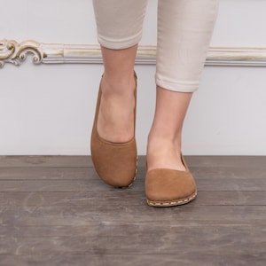 May include: A pair of brown leather ballet flats with a stitched edge detail. The flats are on a wooden floor.