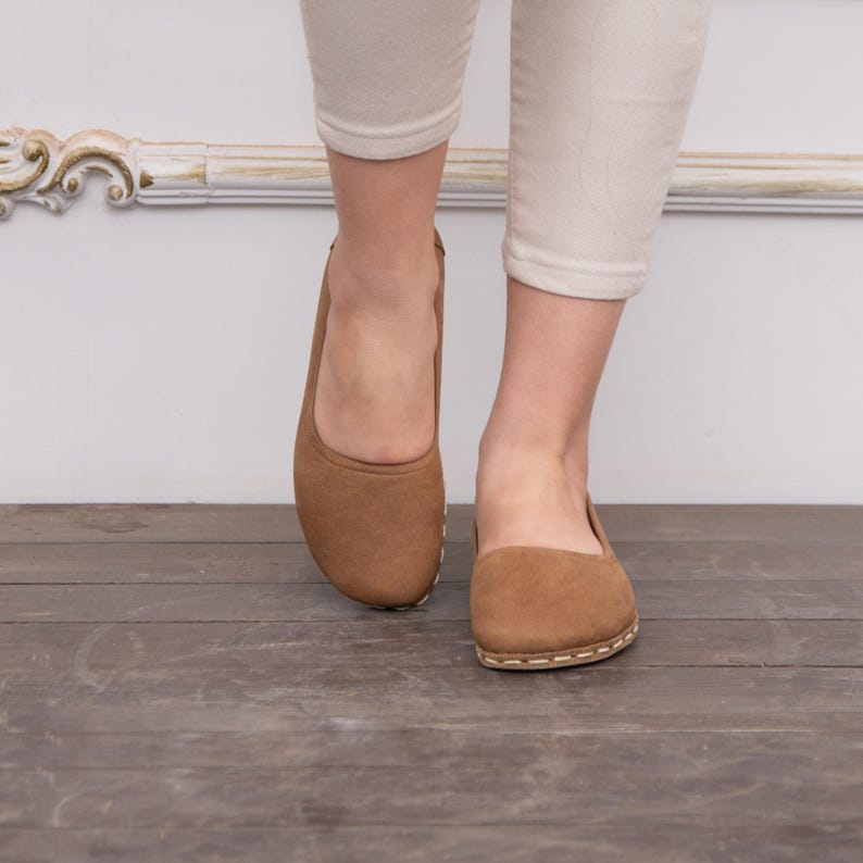May include: A pair of brown leather ballet flats with a stitched edge detail. The flats are on a wooden floor.