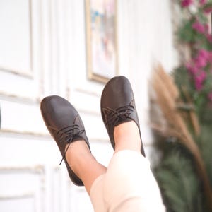 Puede incluir: Un par de zapatos de cuero marrón oscuro con cordones. Los zapatos se muestran desde abajo, con los pies y los tobillos visibles. Los zapatos tienen un diseño clásico y parecen nuevos.