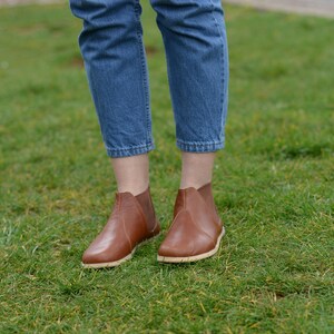 May include: A pair of brown leather ankle boots with a slightly rounded toe and a pull-on design. The boots are worn by a person wearing blue jeans.