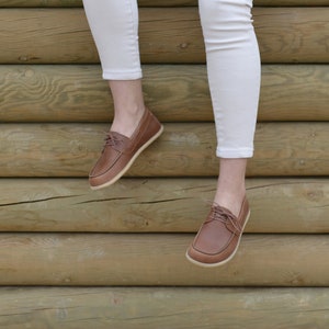 May include: Brown leather boat shoes with laces. The shoes are worn by a person wearing white pants and are photographed against a wooden background.