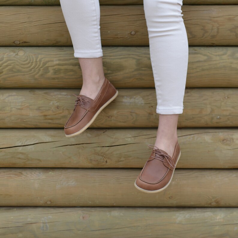 May include: Brown leather boat shoes with laces. The shoes are worn by a person wearing white pants and are photographed against a wooden background.