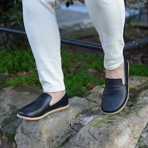 May include: Black leather slip-on shoes with a tan sole. The shoes are worn with off-white pants. The image is taken outdoors on a stone surface.
