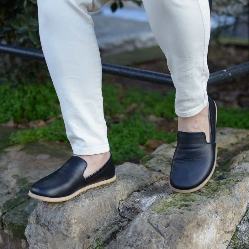 May include: Black leather slip-on shoes with a tan sole. The shoes are worn with off-white pants. The image is taken outdoors on a stone surface.