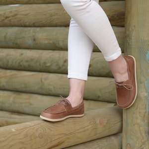 May include: A pair of brown leather boat shoes with laces. The shoes are worn by a person wearing white pants and standing on a wooden surface.