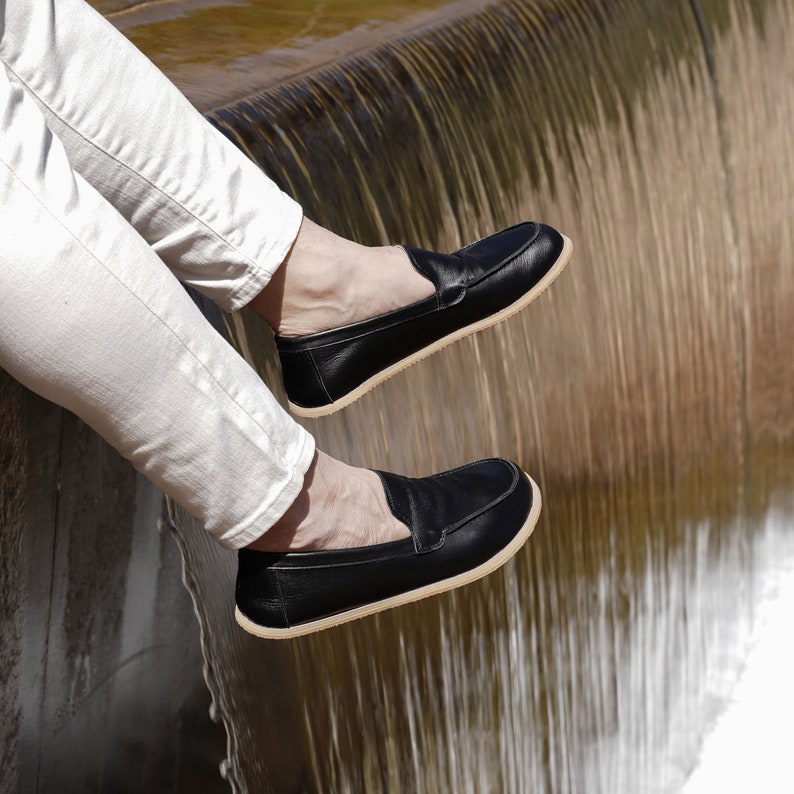 Op de afbeelding: Een paar zwarte leren loafers met een cr&egrave;mekleurige zool. De loafers worden gedragen door een persoon die op een rand zit met een waterval erachter.