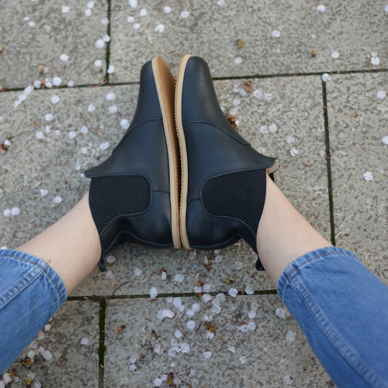May include: A pair of black ankle boots with elastic side panels and a light brown sole. The boots are made of a smooth material and are shown from a top-down perspective. The boots are resting on a gray surface with white flower petals scattered around.