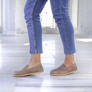 May include: A pair of taupe suede loafers with a rounded toe and a light-colored sole. The shoes are worn with blue denim jeans. The loafers have a classic, slip-on design.