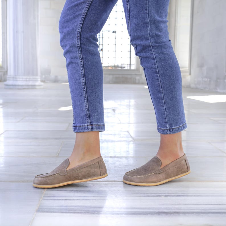 May include: A pair of taupe suede loafers with a rounded toe and a light-colored sole. The shoes are worn with blue denim jeans. The loafers have a classic, slip-on design.