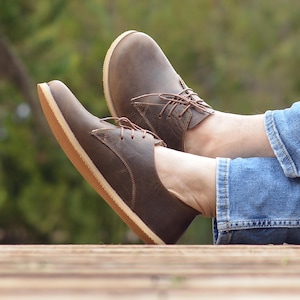Op de afbeelding: Een paar bruine leren veterschoenen met een crèmekleurige zool. De schoenen worden gedragen door iemand die een blauwe jeans draagt.