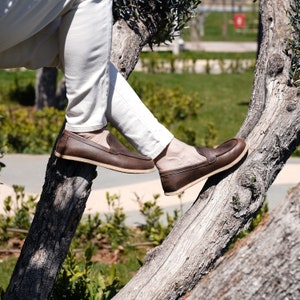 May include: A person wearing brown leather loafers with a slip-on design, sitting on a tree branch.