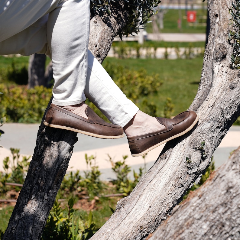May include: A person wearing brown leather loafers with a slip-on design, sitting on a tree branch.