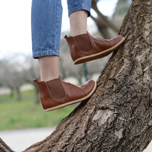 May include: A person wearing brown leather ankle boots with elastic side panels and a pull-on tab. The boots are worn with blue jeans and the person is standing on a tree trunk.