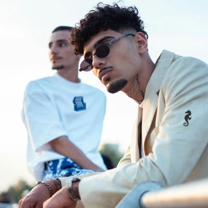 Puede incluir: Dos hombres con gafas de sol, uno con una camiseta blanca y el otro con una chaqueta beige con un caballito de mar negro bordado en la manga.