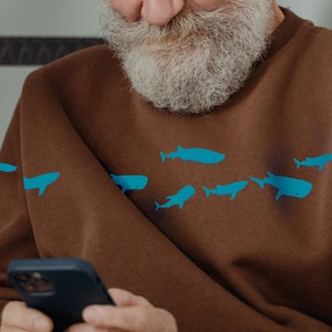 May include: A man wearing a brown sweatshirt with a blue whale and fish graphic print. He is holding a black smartphone in his hands.